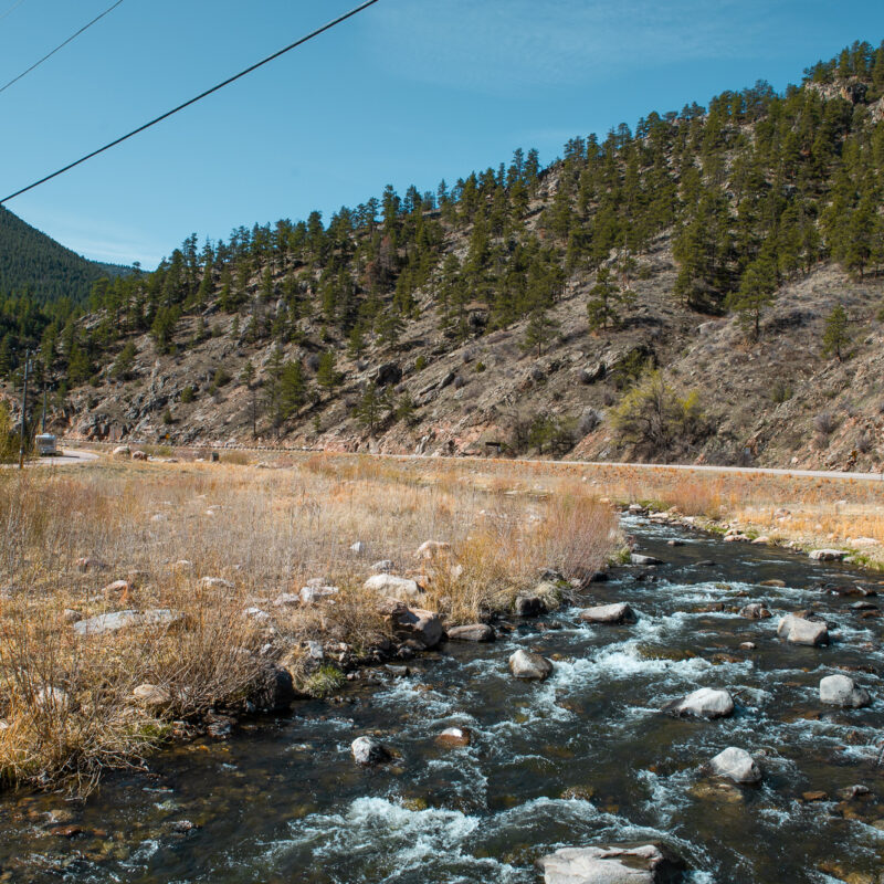 Rocky stream, grassy field, pine-covered mountains, blue sky, power lines.