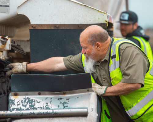 Worker in yellow vest inspects outdoor machinery; another stands behind.