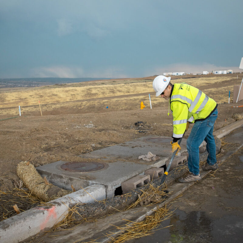 Worker in safety gear checks storm drain beside rural road.