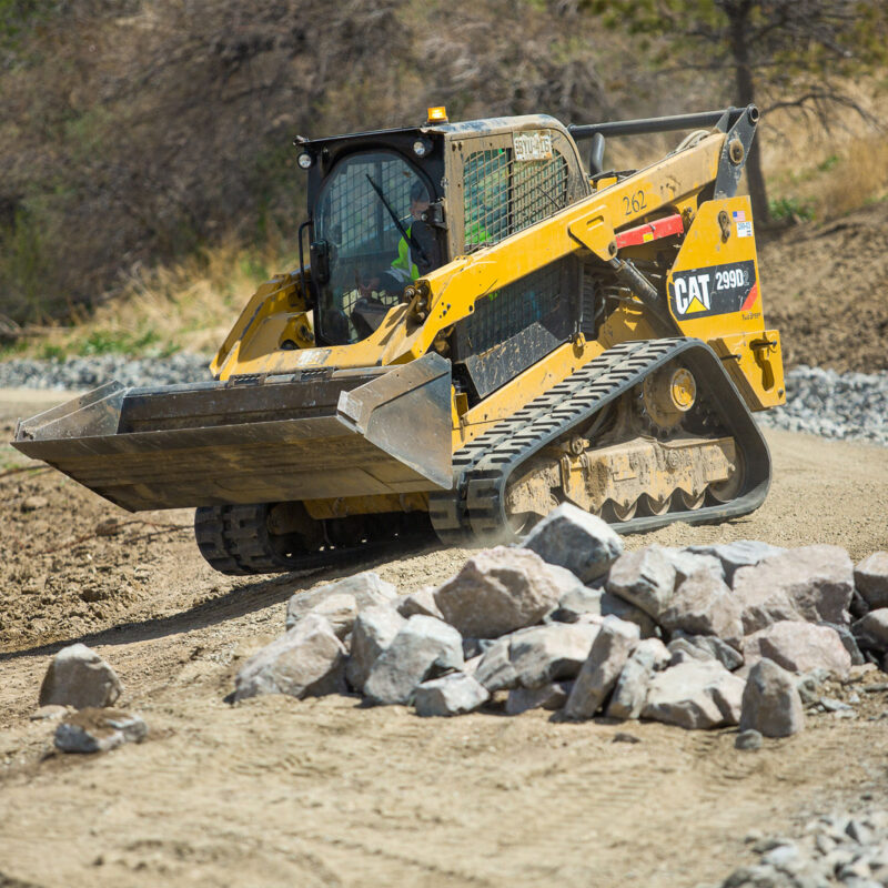 Yellow skid steer loader moves dirt near rocks on a path.
