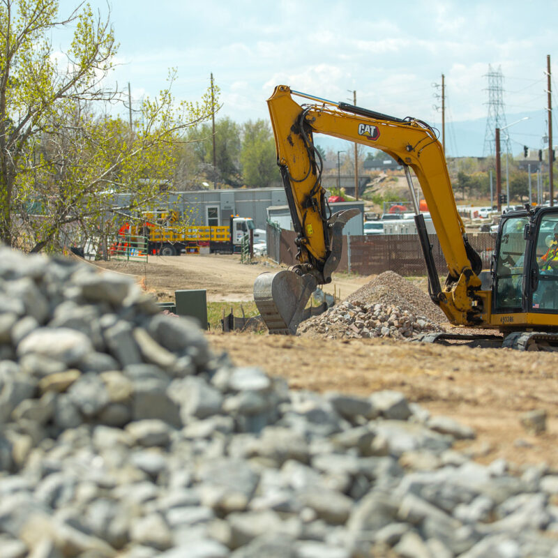 Yellow excavator moving dirt, rocks, with workers in background.