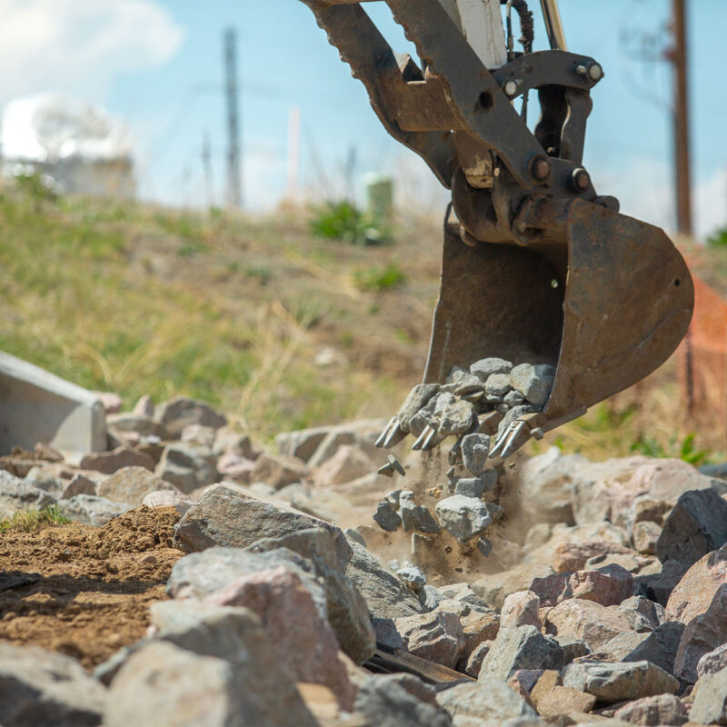 Excavator bucket lifts rocks amid dirt at construction site.