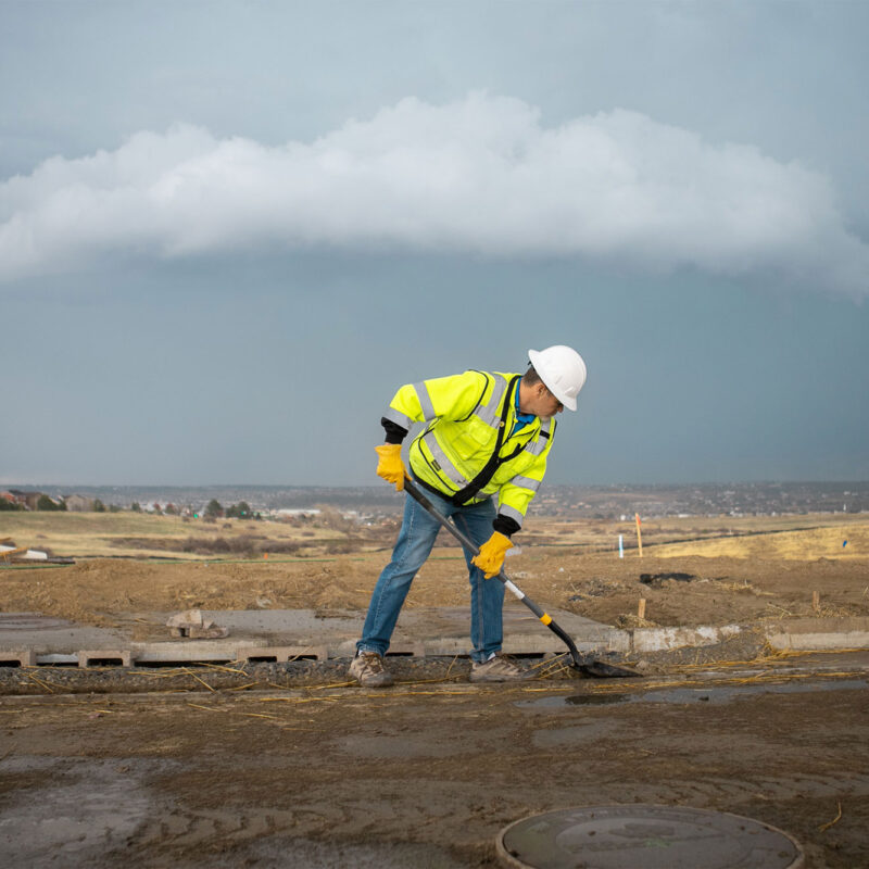 Worker in safety gear shovels on rural road under clouds.