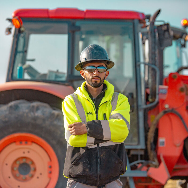 Worker in hard hat and vest stands before red tractor.