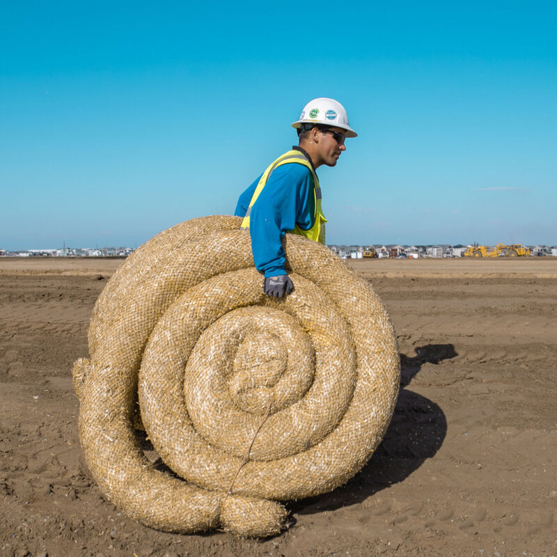 Worker in helmet and vest carries straw mat on dirt site.