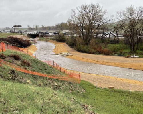 Creek through grass by a highway, orange fencing on banks.