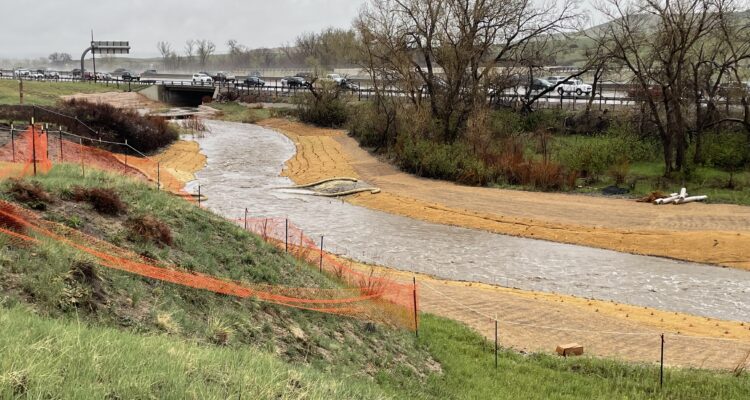 Creek through grass by a highway, orange fencing on banks.