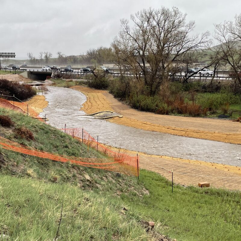 Creek through grass by a highway, orange fencing on banks.