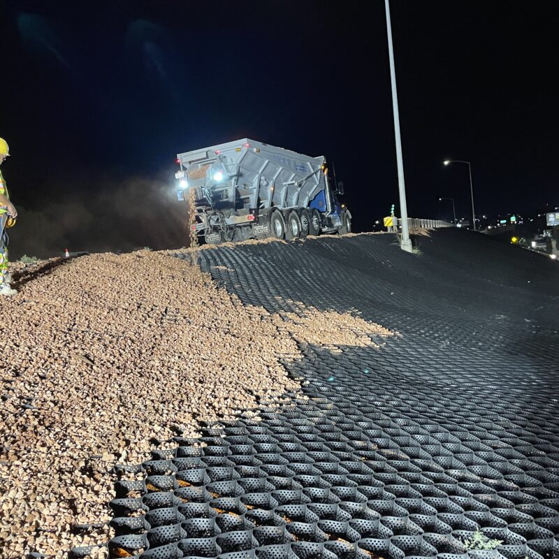 Worker watches dump truck spread gravel on geocell grid at night.