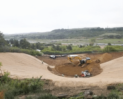 Excavators and workers dig a large earth pit near greenery.