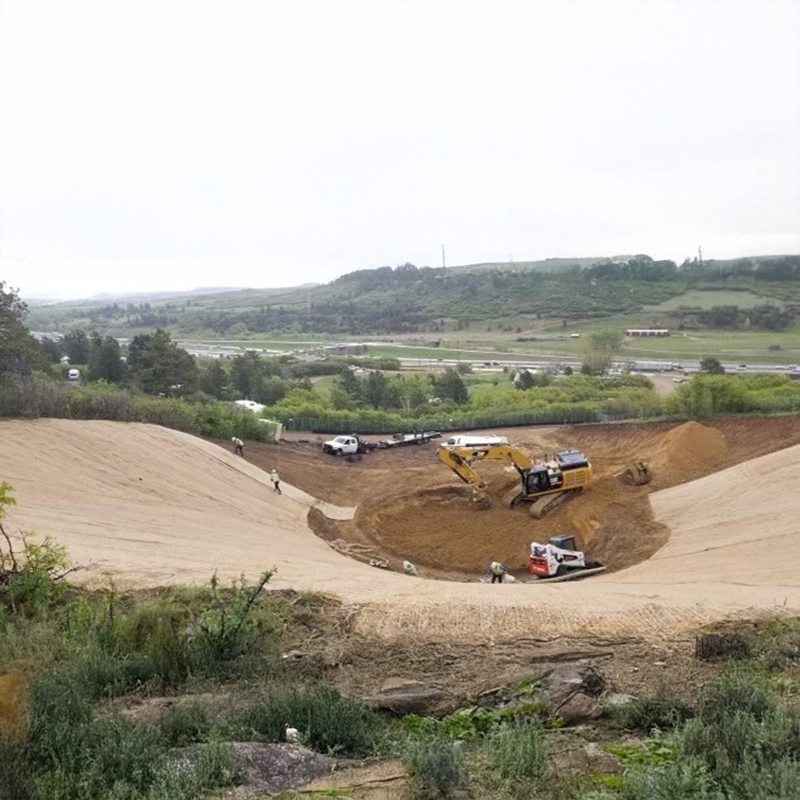 Excavators and workers dig a large earth pit near greenery.
