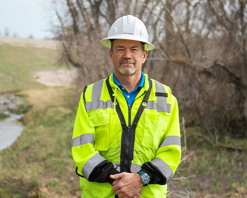 Man in white hard hat and yellow safety jacket outdoors.