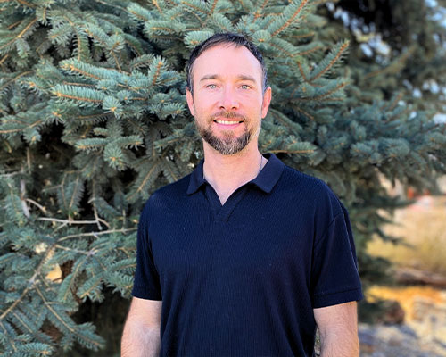 Man in black polo smiles and stands before a pine tree.