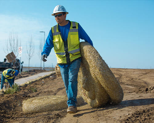 Worker in safety gear carries wire mesh at construction site.