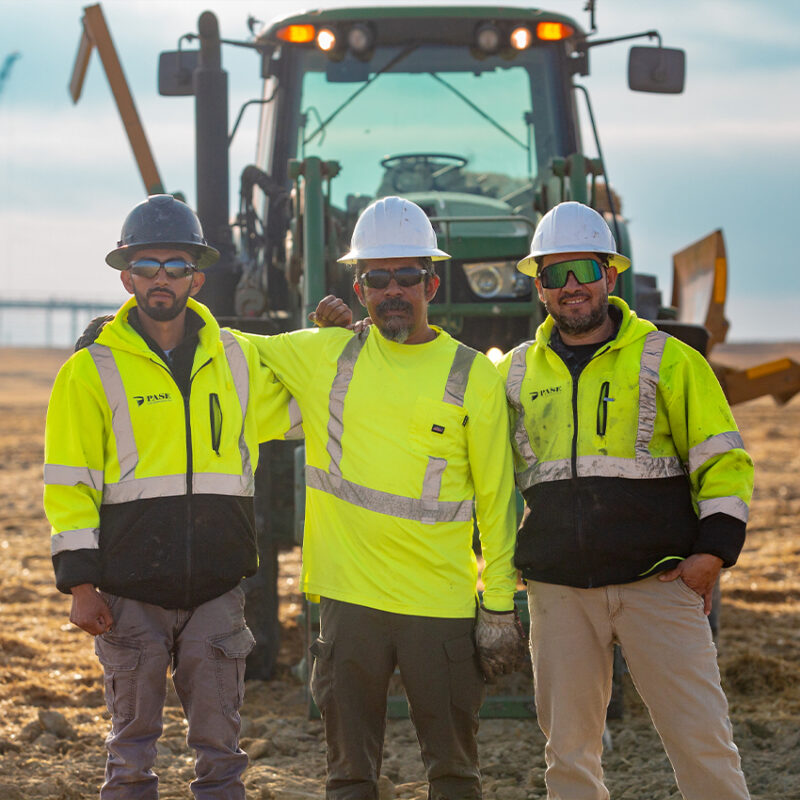 Three workers in safety gear pose by tractor at site.
