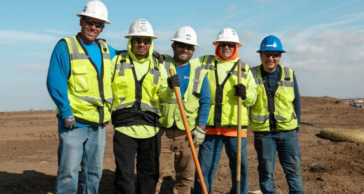 Five construction workers with vests and helmets on sunny worksite.