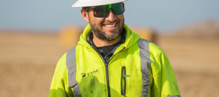 Man in white hard hat and yellow jacket at worksite.